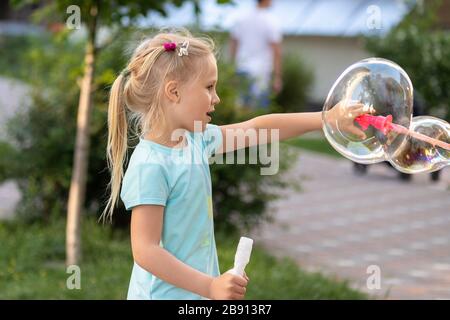 Ritratto di piccola carina ragazza bionda caucasica divertirsi e gioia soffiando grandi bolle di sapone giocando sulla strada della città parco all'aperto. Allegro adorabile bambino Foto Stock