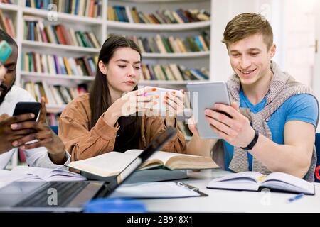 Amici multietnici, studenti universitari stanno studiando in biblioteca e utilizzando i loro gadget, smartphone e tablet, mentre hanno pausa durante la preparazione per Foto Stock