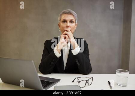 Ritratto minimo di donna d'affari maturo elegante che guarda via con decisione mentre si siede contro il muro grigio sul posto di lavoro, spazio di copia Foto Stock