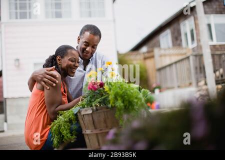 Felice coppia metà-adulto accovacciata accanto ad un cesto di legno pieno di piante e verdure in una strada suburbana. Foto Stock