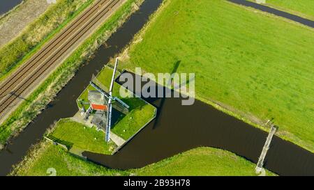 Veduta aerea di un vecchio mulino tradizionale olandese a vento sulla campagna nei Paesi Bassi con una diga, canali, ferrovia, ponte e una strada.