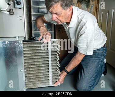 Uomo caucasico senior che cambia un filtro aria sporca piegato nel sistema di fornace HVAC nel seminterrato di casa Foto Stock