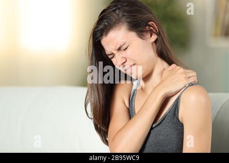 La ragazza lamenta il dolore alle spalle che si siede su un divano nel soggiorno a casa Foto Stock