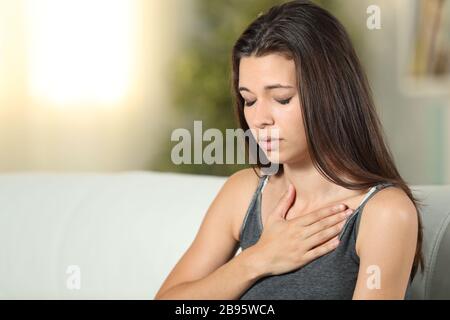 Ragazza con problemi respiratori che tocca il petto seduto su un divano nel soggiorno a casa Foto Stock