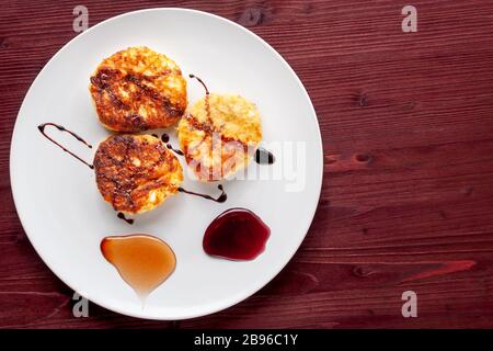 Le frittelle di formaggio casolare su un piatto sono decorate con miele con mirtilli, miele con fragole e sciroppo di carruba. Colazione sana e dietetica. Foto Stock