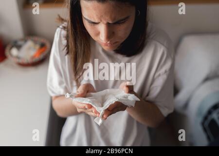 Giovane donna malata starnutita in un tessuto mentre ricopriva con un trapunta blu in casa sua Foto Stock