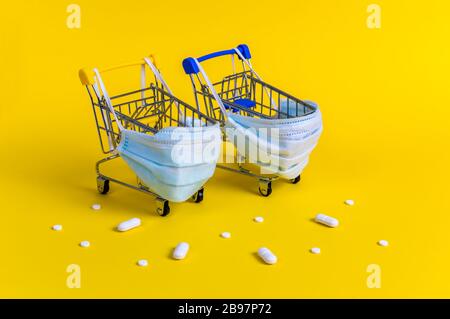 Due carrelli con maschere mediche per la protezione antivirus circondati da pillole bianche su sfondo giallo. Concetto creativo di shopping online sicuro Foto Stock