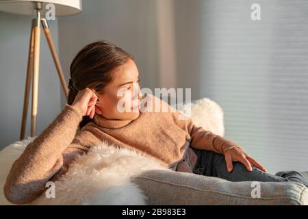 Soggiorno casa asiatica donna rilassante pensivo guardando fuori la finestra sedersi nel divano del soggiorno godendo del tempo libero all'interno durante focolaio di coronavirus Foto Stock