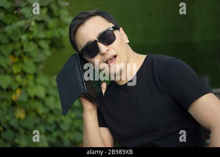 Giovane asiatico con t-shirt nera e occhiali da sole che parla con lo smartphone Foto Stock