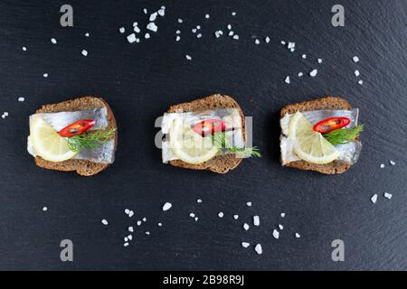 Panini con aringhe sottaceto al limone, peperoncino e un rametto di aneto. Cibo sano. Sfondo nero. Foto Stock