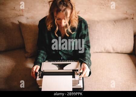 Giovane scrittrice femminile che usa la macchina da scrivere mentre si siede sul divano a casa Foto Stock