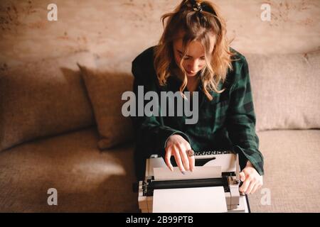 Giovane scrittrice femminile che usa la macchina da scrivere mentre si siede sul divano a casa Foto Stock