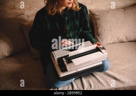 Giovane scrittrice femminile che usa la macchina da scrivere mentre si siede sul divano a casa Foto Stock