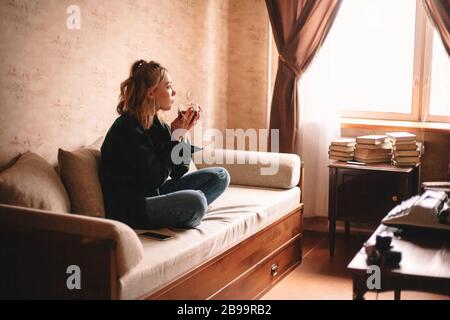 Giovane donna premurosa bere tè e mangiare cioccolato guardando attraverso la finestra seduta sul divano in soggiorno a casa Foto Stock