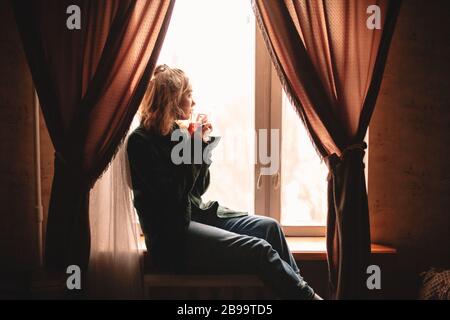 Giovane donna premurosa che beve il tè e guarda attraverso la finestra mentre si siede sul davanzale a casa Foto Stock
