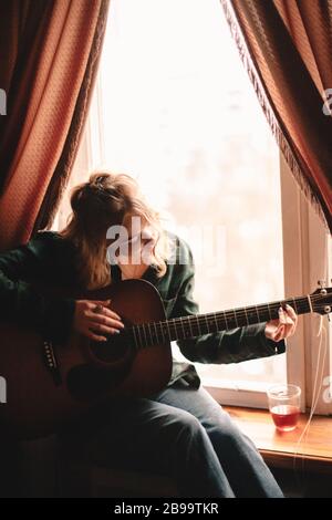 Giovane donna che suona la chitarra mentre si siede su windowsill a casa Foto Stock