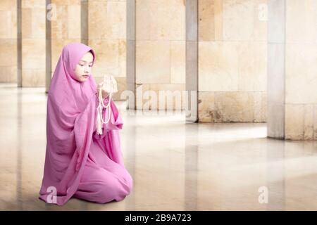 Donna musulmana asiatica in velo seduta e pregando con perline di preghiera sulle sue mani all'interno della moschea Foto Stock