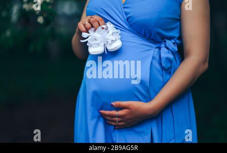 donna incinta che tiene il bootee bianco del bambino che indossa in abito blu sullo sfondo del parco naturale al tramonto. Foto Stock