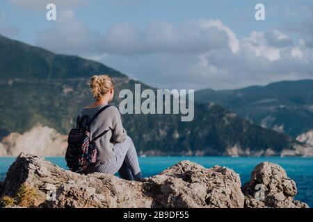 Donna con zaino colorato che si trova sulla costa rocciosa guardando al mare e l'isola all'orizzonte. Tempo tempestoso. Foto Stock