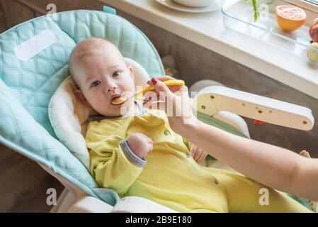 La mamma alimenta la purea di verdure del bambino in un seggiolone. La madre mette un cucchiaio di schiacciamento nella bocca del bambino. Il bambino mangia cibo. Foto Stock