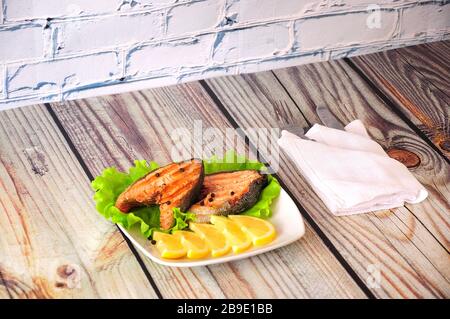 Due bistecche di salmone rosa alla griglia giacciono su un piatto di ceramica bianca decorato con fette di limone e una foglia di lattuga accanto ad un tovagliolo bianco con posate, il wh Foto Stock
