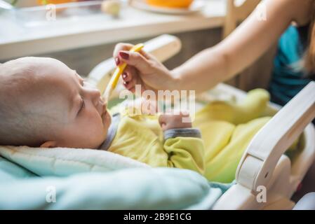 La mamma alimenta la purea di verdure del bambino in un seggiolone. La madre mette un cucchiaio di schiacciamento nella bocca del bambino. Il bambino mangia cibo. Foto Stock