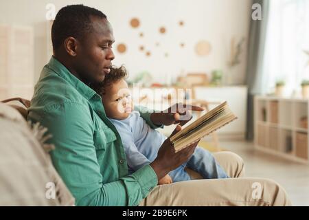 Vista laterale ritratto di cura afro-americano papà lettura libro a cute baby figlio a casa, copia spazio Foto Stock