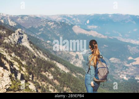 Giovane bella ragazza viaggia da sola in montagna in primavera o autunno, guarda in lontananza e gode di natura, rocce e foreste verdi, vista del paesaggio. Uno zaino dietro e abbigliamento sportivo, libertà e leggerezza Foto Stock