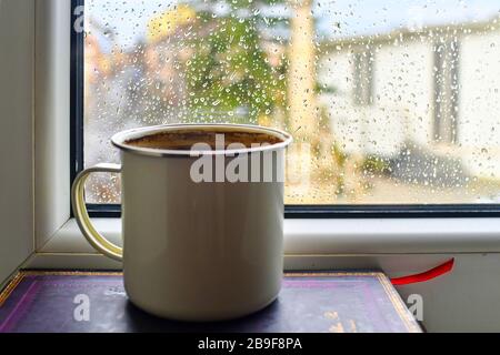 Tazza di caffè sullo sfondo della finestra piovosa. Tazza di metallo su mucchio di libri. Tradizionale caffè ceco - caffè turco Foto Stock