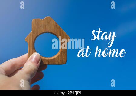 Piccola casa giocattolo in legno in mano su sfondo blu cielo con parole rimanere a casa. Concetto di quarantena. Foto Stock