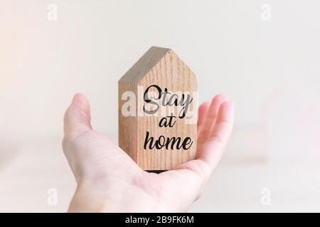 Piccola casa giocattolo in legno in mano su sfondo bianco pastello con parole rimanere a casa. Concetto di quarantena. Foto Stock