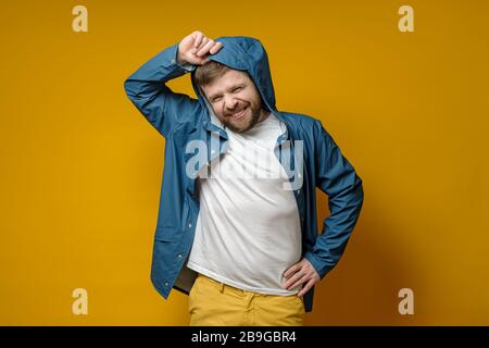 Attraente uomo caucasico bearded in un impermeabile mette su un cappuccio e sorride carino. Isolato su uno sfondo giallo. Foto Stock