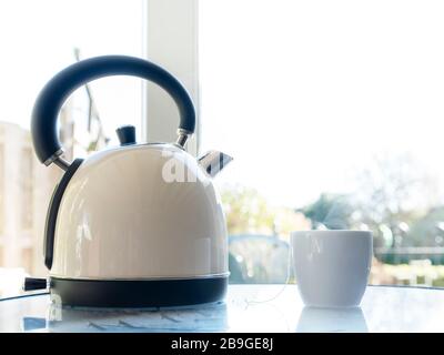 bollitore elettrico con acqua bollente per il tè alle erbe su un tble davanti ad una finestra. Concentrato selettivo sul bollitore e sulla tazza da tè Foto Stock