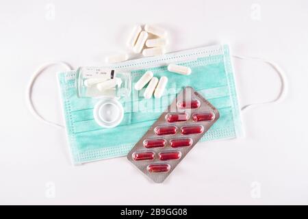 Capsule bianche di medicina che fuoriescono dal flacone di vetro accanto a un blister con pillole rosse sulla parte superiore di una maschera chirurgica (vista dall'alto su sfondo bianco) Foto Stock