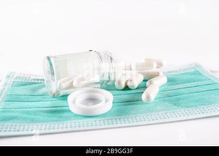 Capsule bianche di medicina che fuoriescono dal flacone di vetro sulla parte superiore di una maschera chirurgica (vista dell'occhio della rana su sfondo bianco) Foto Stock