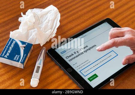Una donna che utilizza un ipad per controllare il sito web NHS per consigli sui simpton Coronavirus Covid-19. Con paracetamolo, tessuto e termometro. Foto Stock