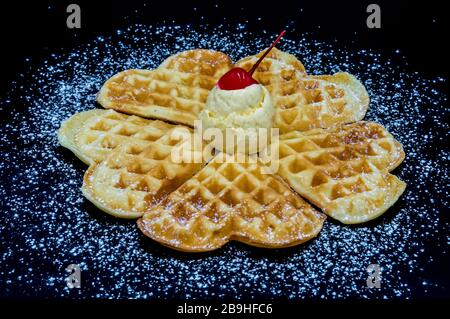 Waffle fatti in casa con zucchero in polvere, un cucchiaio di gelato alla vaniglia e una ciliegia su uno sfondo nero, immagine isolata del cibo. Foto Stock