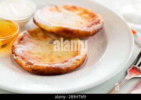 Cheesecake ucraini colazione con miele e crema su piatto bianco. Frittelle o frittelle di formaggio casolare Foto Stock