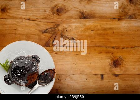 Un singolo pan di Spagna al cioccolato, con una salsa appiccicosa al cioccolato, su un piatto bianco con un cucchiaio, girato su uno sfondo di legno con spazio per la copia Foto Stock