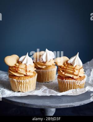 Cupcake di carote con noci e spezie decorate con biscotti a forma di cuore, meringa e caramello salato su carta bianca e sfondo scuro. Spazio libero Foto Stock