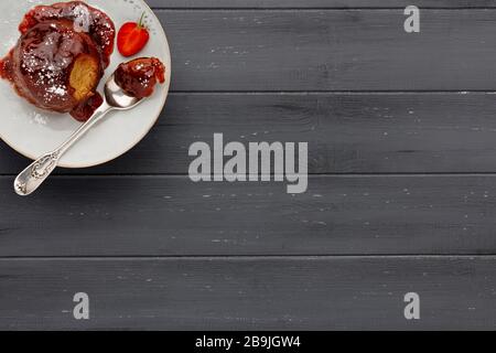 Un singolo pan di Spagna di fragola, con una salsa di fragole appiccicosa, su un piatto bianco con un cucchiaio, su una superficie di legno grigio scuro con spazio per la copia Foto Stock