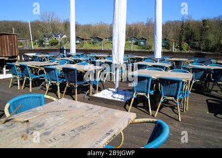 Viersen (Hariksee), Germania - Marzo 22. 2020: Vista sulla terrazza vuota del ristorante chiuso con sedie e tavoli al lago durante il giorno di sole durante il forte arredo Foto Stock