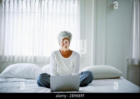 Donna matura nei suoi pigiami siede a gambe incrociate su un letto e guarda verso il basso un notebook. Foto Stock