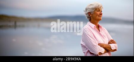 Content donna matura con le sue braccia incrociate si erge su una spiaggia e si affaccia sul mare. Foto Stock