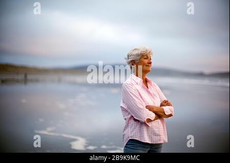 Content donna matura con le sue braccia incrociate si erge su una spiaggia e si affaccia sul mare. Foto Stock