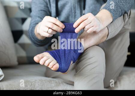 L'uomo si dà il primo aiuto che arrotola nastro di bendaggio blu sopra il piede Foto Stock