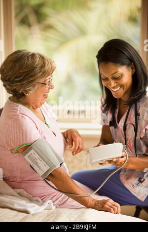 L'infermiera femminile utilizza un bracciale per la misurazione della pressione sanguigna e un monitor per la misurazione della pressione su una donna anziana che siede sul letto. Foto Stock