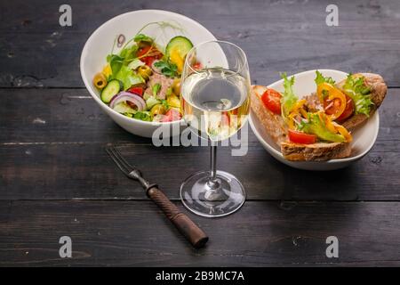 Cibo ancora vita. Vino bianco in un bicchiere, insalata con verdure e microgreens, tartine con paté e pepe dolce su un tavolo di legno scuro. Foto Stock