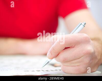 Primo piano di una mano d'uomo che tiene una matita che risolve i crosswords che portano una camicia rossa. Foto Stock