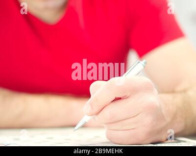 Primo piano di una mano d'uomo che tiene una matita che risolve i crosswords che portano una camicia rossa. Foto Stock